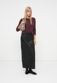 Γυναίκα φορώντας ένα αφράτο σκούρο μωβ πουλόβερ και μακριά μαύρη δερμάτινη φούστα, κρατώντας τσάντα ώμου με σχέδιο από δέρμα φιδιού, standing against a white background.