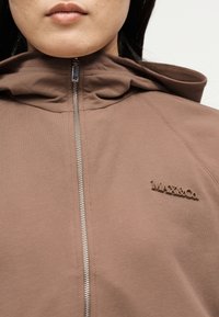 Γυναίκα που φοράει καφέ φούτερ με φερμουάρ και ανάγλυφο λογότυπο "MAX&Co." στο στήθος, φαίνεται από το πηγούνι μέχρι το πάνω μέρος του στήθους.