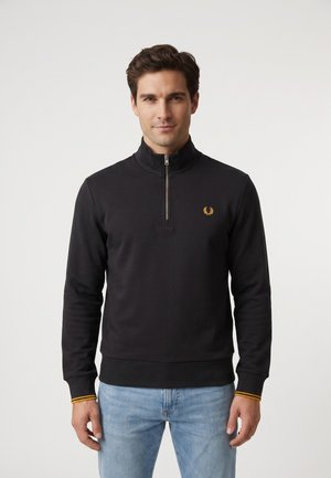 Uomo che indossa un maglione nero con zip corta, con un piccolo logo dorato sul petto e jeans azzurri su sfondo uniforme.