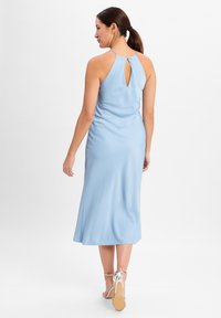 Hellblaues ärmelloses Kleid mit einem Schlüsselloch-Ausschnitt im Rücken, glatter Textur und einer fließenden Silhouette, die bis zur Mitte der Waden reicht. Sichtbare silberne Riemchen-Highheels.