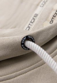 Tissu beige avec un cordon blanc passé à travers un œillet en métal noir, imprimé "ombre wear" et une étiquette de tissu "ombre" visible en arrière-plan.