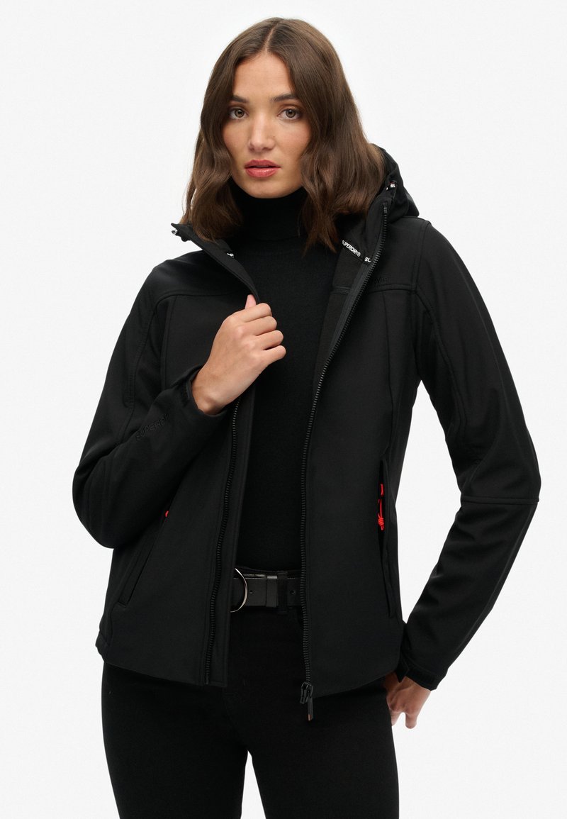 Vrouw met schouderlang bruin haar, gekleed in een zwarte zip-up jas over een zwarte coltrui, staande voor een effen witte achtergrond.