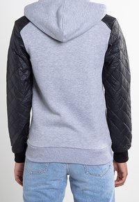 Sudadera gris con un diseño acolchado de cuero negro en los hombros y los brazos. Textura suave, con un bolsillo tipo canguro y puños acanalados.