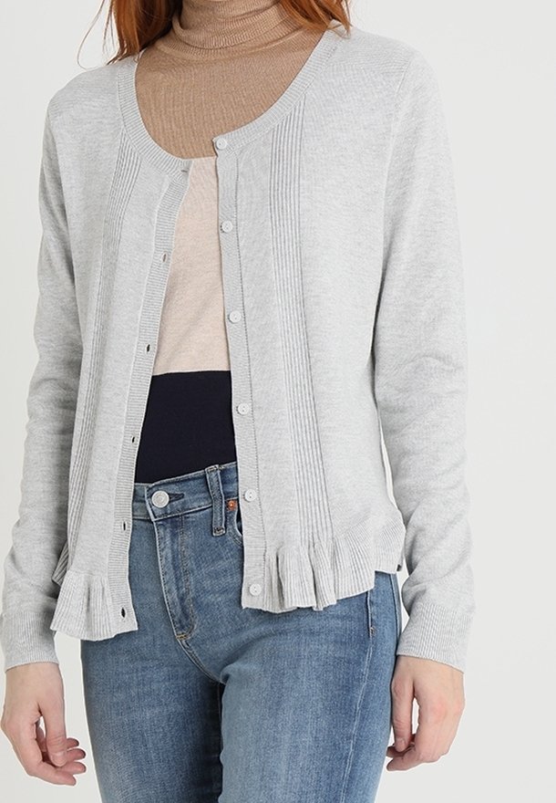 Femme portant un cardigan gris clair boutonné avec des détails côtelés et un ourlet à volants, sur un pull à col roulé beige et un jean bleu.