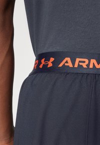 Navy athletic shorts with an elastic waistband featuring a contrasting orange "UNDER ARMOUR" logo and smooth texture. Close-up view.