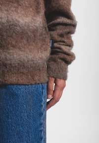Maglione marrone lavorato a maglia con texture morbida, caratterizzato da un polsino arrotolato, abbinato a denim blu. Etichetta visibile sul lato sinistro del maglione.
