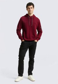 Bordeauxrode hoodie met een voorzak, capuchon met trekkoord en een ritszak op de mouw. Gecombineerd met een zwarte broek en witte sneakers.