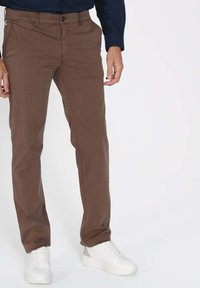 Chinos marron en coton au toucher lisse, avec une fermeture éclair et des poches latérales, associés à des baskets blanches.