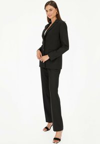Czarny garnitur szyty na miarę, składający się z jednorzędowej marynarki z ostrymi klapami, dopasowanych spodni oraz gładkiej tkaniny. Całość dopełniają szpilki.