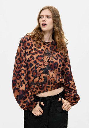 Kvinde med langt hår iført en leopardmønstret sweater med Daisy Duck-motiv, ser overrasket ud, hænderne i lommerne på sorte bukser.