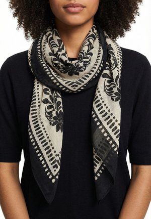 Femme portant une écharpe beige clair avec des motifs floraux et géométriques noirs sur un haut noir à manches courtes, laissant apparaître le cou et les épaules.