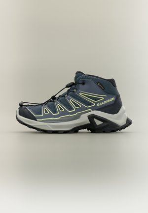 Tumši zila un pelēka vidēja augstuma pārgājienu apava ar dzelteniem akcentiem, ātrās auklas sistēmu, Salomon logotipu un robainu melnu zoli.