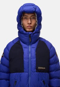 Giacca a vento blu con colletto alto e cappuccio integrato. Presenta dettagli blu navy, due tasche con zip e un patch con logo sul petto.