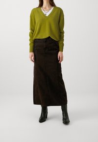 Suéter verde de escote en V combinado con una falda maxi de denim marrón. El conjunto se complementa con botas de tacón negras y joyería de plata en capas.