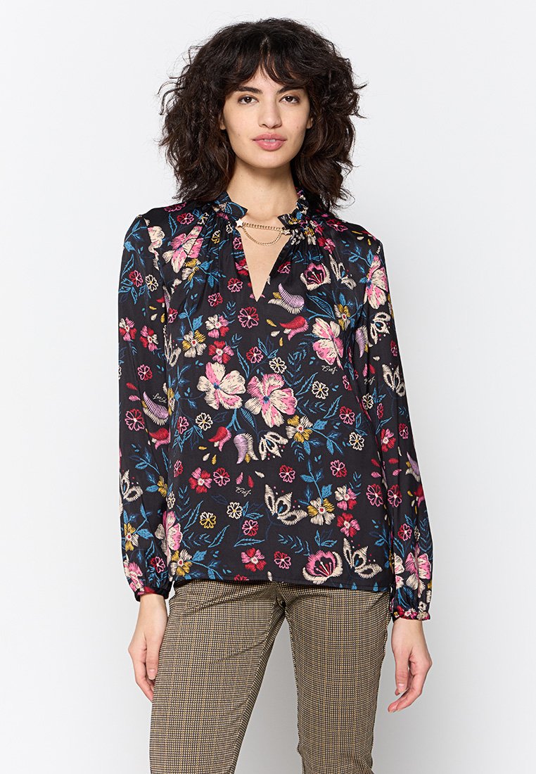 Blusa nera con motivi floreali colorati, maniche lunghe, scollatura a V e arricciature sulle spalle. Abbinata a pantaloni marroni con motivi.