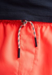 Shorts de bain orange vif avec un cordon de serrage rayé noir et blanc. Un ourlet supérieur bleu marine foncé est visible au-dessus de la ceinture.