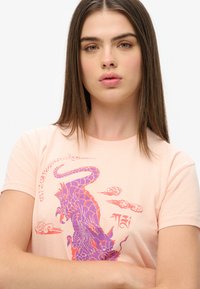 Camiseta de algodón de color rosa claro con un gráfico de dragón púrpura y acentos de nubes. Mangas cortas y ajuste clásico.