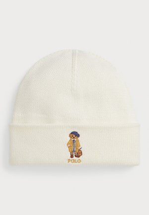 Bonnet en tricot crème avec un revers, mettant en avant un ours brodé portant un manteau et un chapeau, ainsi que le texte "Polo" en dessous du design.