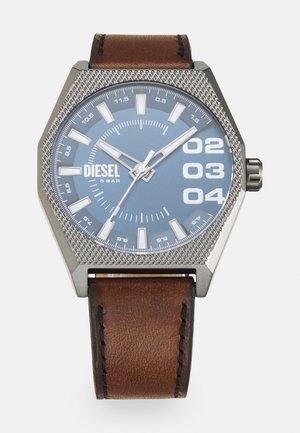 Montre argentée hexagonale avec cadran bleu, index horaires blancs et bracelet en cuir marron. Présente une lunette texturée et une inscription de logo.