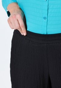 Zwarte elastische broek met textuur, gecombineerd met een lichtblauw kreukelig shirt met een knoopsluiting en korte mouwen.