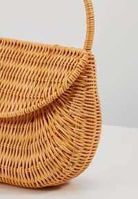 Petit sac à main en rotin tressé avec rabat incurvé et forme arrondie, présentant une seule poignée sur un fond clair uni.