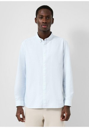 Młody mężczyzna w jasnoniebieskiej koszuli na guziki i beżowych spodniach, stojący na tle jednolitego szarego tła, zwrócony przodem.