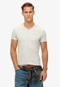 Lys grå bomulds T-shirt med V-udskæring, korte ærmer, parret med blå jeans og en ternet skjorte bundet om livet. Har et diskret logo på brystet.