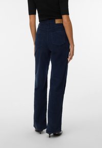 Femme portant un pantalon en velours côtelé bleu foncé taille haute et un haut noir, debout, vue de dos sur un fond blanc uni.