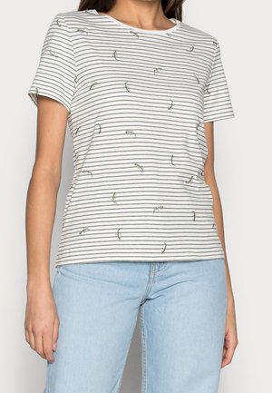 Kvinde iført en hvid kortærmet skjorte med tynde sorte striber og små firkantede print, kombineret med lyseblå jeans.