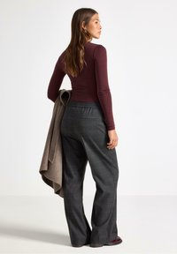 Kvinde med langt hår iført en maroon langærmet bluse og grå ruted bukser med vide ben, som holder en sammenfoldet beige frakke over armen.