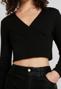 Schwarzes, geripptes Langarmoberteil mit V-Ausschnitt und Wickeldesign; kurze Länge. Kombiniert mit hochgeschnittenen schwarzen Lederhosen mit Knopfdetails.