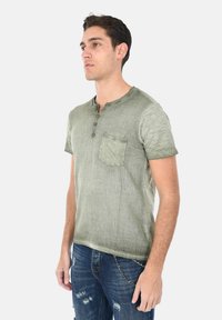 Maglietta henley a maniche corte verde oliva con taschino sul petto, apertura con tre bottoni e tessuto texturizzato; abbinata a jeans blu strappati.