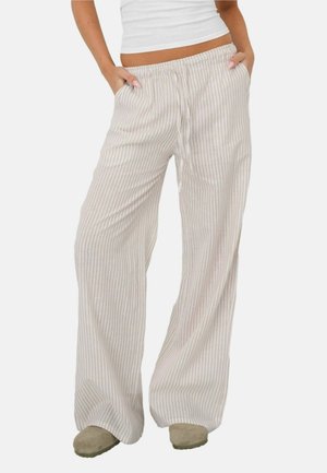 Frau trägt beige-weiß gestreifte, weit geschnittene Hose mit vertikalen Streifen, Hände in den Taschen, kombiniert mit weißem Crop-Top und beigen Schuhen.