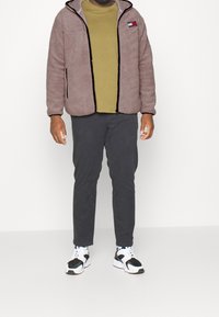 Fleecejakke i lysebrun med sort kant, med hætte og brystlogo. Kombineret med en grøn top og mørkegrå bukser, afsluttet med sorte og hvide sneakers.