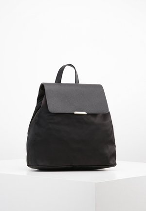 Sac à dos noir avec rabat texturé et poignée supérieure, présenté en position verticale sur un piédestal blanc contre un fond blanc uni.