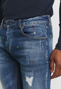 Jeans de mezclilla de color azul desgastado, con detalles desgastados y manchas de pintura en rojo, verde y blanco. Presentan un diseño clásico de cinco bolsillos.