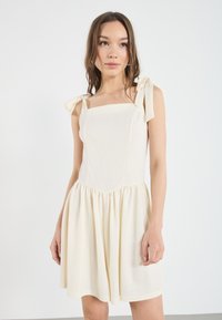 Lichtbeige jurk met vierkante halslijn, geplooide rok en strikbandjes. Gemaakt van een zachte stof, met een gladde textuur en een eenvoudig ontwerp.
