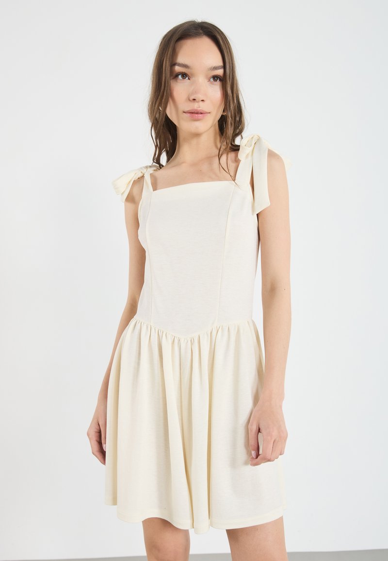 Lichtbeige jurk met vierkante halslijn, geplooide rok en strikbandjes. Gemaakt van een zachte stof, met een gladde textuur en een eenvoudig ontwerp.