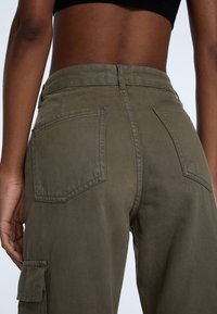 Pantalon cargo en coton vert olive, avec poches arrière et coupe droite. Coutures apparentes et détails de tissu texturé.