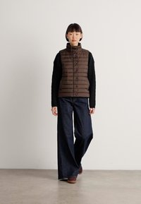 Kvinne iført en brun quiltet vest, svart langermet genser, vide mørke jeans og brune sko som går på et enkelt gulv mot en hvit vegg.