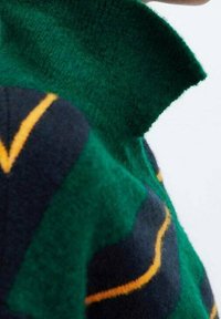 Zielony dziany sweter z wysokim kołnierzem, ciemnoniebieskimi i żółtymi paskami oraz miękką teksturą. Zbliżenie na detal kołnierza.