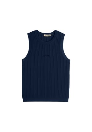 Nuori mies, jolla on yllään tummansininen hihaton ribbineulepaita, harmaat neuloshousut ja beige lippalakki, jossa on tumma logo, seisoo neutraalisti.