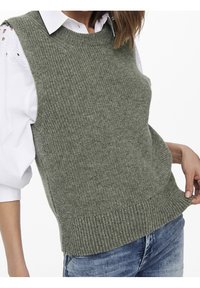 Pull vert sans manches en maille avec une finition texturée, col rond et ourlet côtelé, porté par-dessus une chemise blanche à col.