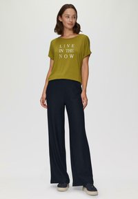 Olijfgroene t-shirt met korte mouwen en witte tekst "LIVE IN THE NOW," gecombineerd met donkere wijde broek en zwarte sneakers.