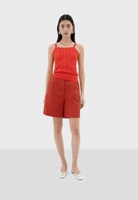 Débardeur rouge côtelé avec décolleté carré, associé à un short plissé couleur rouille. Le modèle porte des chaussures plates argentées. Design simple et épuré.