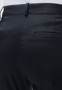 Gros plan sur un pantalon formel noir montrant la ceinture, les passants de ceinture et la poche arrière avec une chemise blanche rentrée.