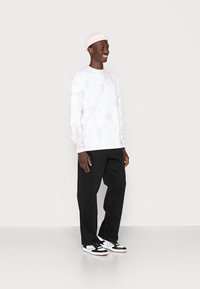 White long-sleeve shirt with pink tie-dye pattern, black relaxed-fit jeans, and black/white sneakers. Pink beanie on head.