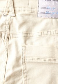 Denim w kolorze off-white z widocznym przeszyciem, boczną kieszenią oraz metką z tekstem i zdobniczymi akcentami na górze.