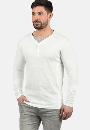 Hvid langærmet henley-skjorte med V-hals og tre-knaps placket, lavet af blødt stof. Parret med lysegrå bukser.