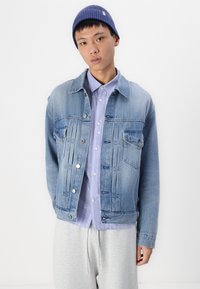 Lichtblauwe denim jas met een klassieke snit, voorzien van twee borstzakken en knoopsluitingen, gedragen over een lichtpaarse gestreepte shirt.